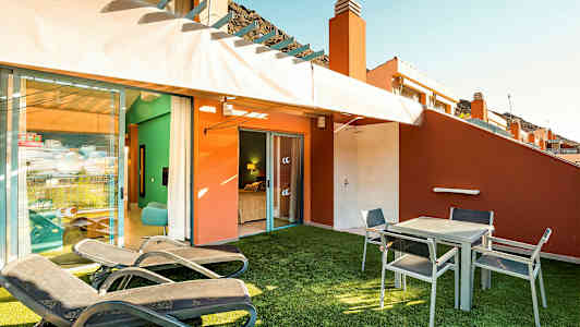 En terrasse med liggestole, bord og stole samt udsigt ind mod et soveværelse. (AI-genereret tekst)