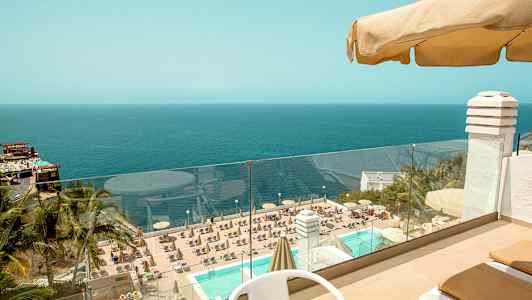 Udsigt fra en terrasse med bord og stole ud mod en pool omgivet af solstole og parasoller, med en udsigt over havet i baggrunden. (AI-genereret tekst)