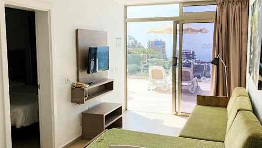 En hotelværelsesudsigt med en grøn sofa og et TV på væggen, med en døråbning til et soveværelse og en terrasse med udsigt over omgivelserne udenfor. (AI-genereret tekst)