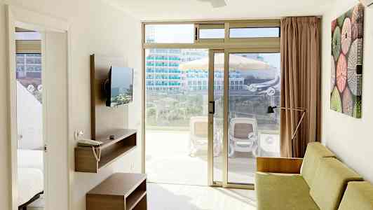 Et lyst hotelværelse med sofa, vægmonteret TV og skydedøre i glas, der fører ud til en møbleret altan. (AI-genereret tekst)