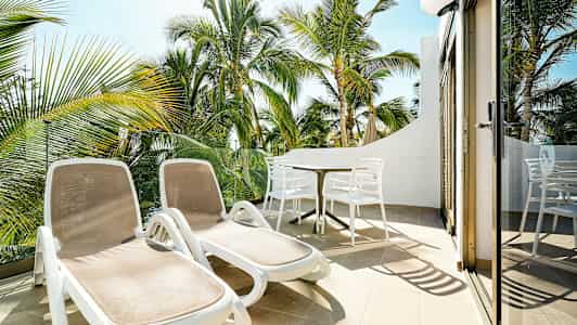 Liggestole og spisebord på en terrasse med udsigt til palmetræer. (AI-genereret tekst)