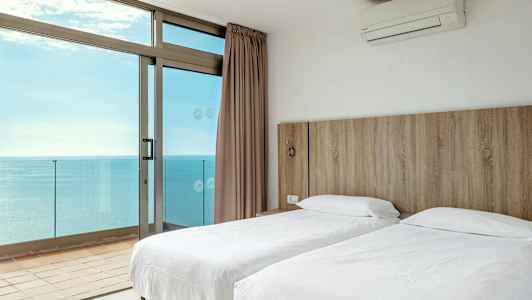 Et hotelværelse med to enkeltsenge og udsigt over havet gennem store vinduer. (AI-genereret tekst)