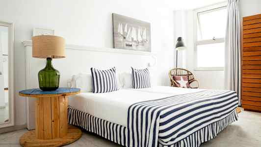 Et soveværelse med en seng beklædt med stribede tekstiler, et bord med en grøn glaslampe og en kurvestol ved siden af. (AI-genereret tekst)