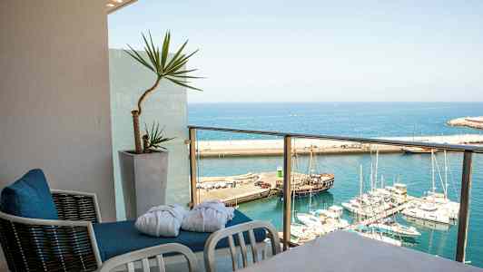 Udsigt over en havn med flere både fra en altan med stole og en potteplante. (AI-genereret tekst)