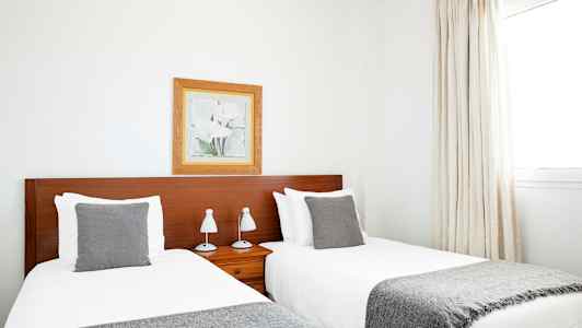 To enkeltsenge med grå tæpper og puder, placeret ved siden af en træfarvet sengegavl. Et indrammet portræt hænger på væggen over sengegavlen. (AI-genereret tekst)