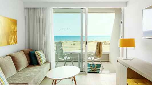 Udsigt fra et lyst hotelværelse med moderne indretning over en solrig strand og havet. (AI-genereret tekst)