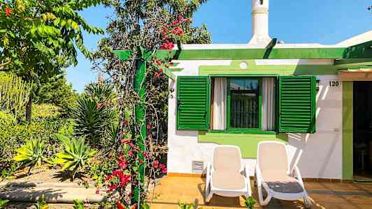 Billede af et hus med grønne skodder og en terrasse med to liggestole. Beplantning med blomster og planter omgiver området. (AI-genereret tekst)