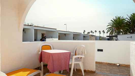 Den viser en terrasse med liggestole og et bord med stole. Baggrunden har små huse og palmer. (AI-genereret tekst)