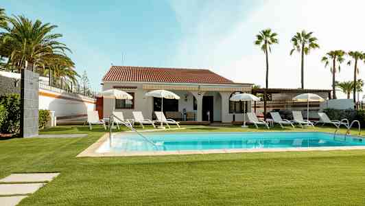 Villa med pool og liggestole, omgivet af græsplæne og palmer. (AI-genereret tekst)
