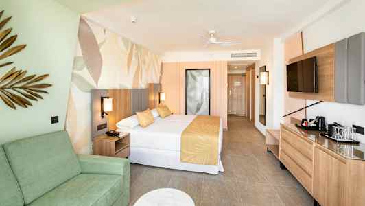 Hotelværelse med seng, sofa og tv. Indretning i lyse farver. (AI-genereret tekst)