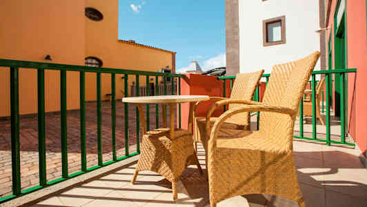 To flettede stole og et lille bord står på en solrig terrasse med farverige bygninger i baggrunden. (AI-genereret tekst)