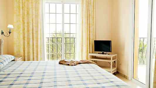 En lys soveværelsesinteriør med en seng, et TV på et bord og store vinduer med gardiner. (AI-genereret tekst)