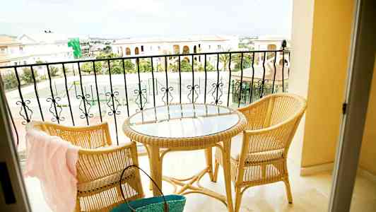 To rottingstole og et rundt bord med glasplade står på en altan med udsigt over stenbygninger og et tennisområde. (AI-genereret tekst)