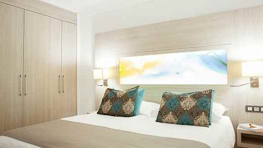 Hotelværelse med en dobbeltseng og dekorative puder, omgivet af lyst træ og lamper. (AI-genereret tekst)