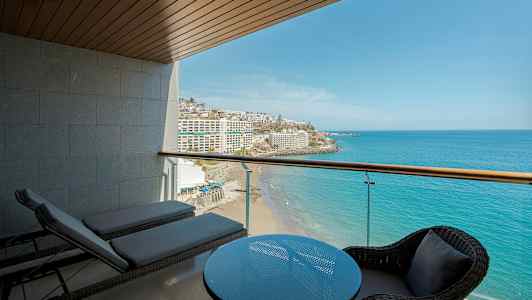 Udsigt fra en altan over havet og en strand, med bord og stole samt solsenge i forgrunden. (AI-genereret tekst)