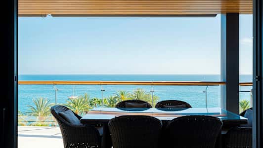 Billede af en terrasse med bord og stole, havudsigt og palmer i baggrunden. (AI-genereret tekst)