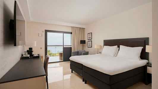 Et hotelværelse med en stor seng, et skrivebord og et TV. Gennem glasdøren ses en balkon og udsigt mod havet. (AI-genereret tekst)