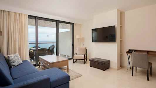 En stue med blå sofa, træbord og stole ved et stort vindue mod altanen. (AI-genereret tekst)