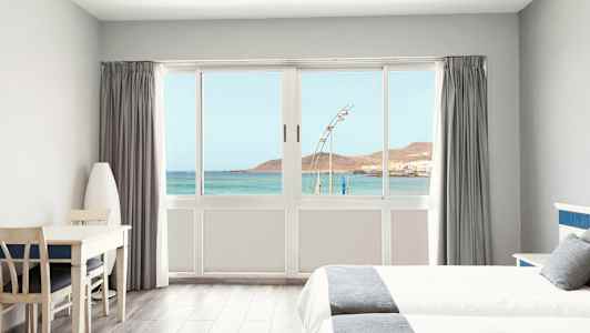 Lyst hotelværelse med udsigt over havet gennem en stor vinduesfront. (AI-genereret tekst)