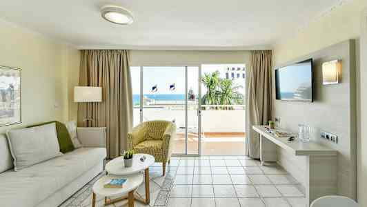 En stue med lyse møbler og en stor glassdør, der fører til en altan med havudsigt. (AI-genereret tekst)