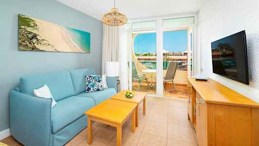 En stue med en blå sofa, to træborde, et fjernsyn på et skab og udsigt til en terrasse med havemøbler. (AI-genereret tekst)