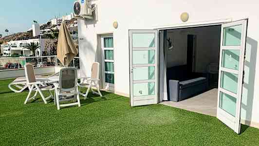 Udendørsområde med grøn græsplæne, siddegruppe og åbne døre til et indendørsområde. (AI-genereret tekst)