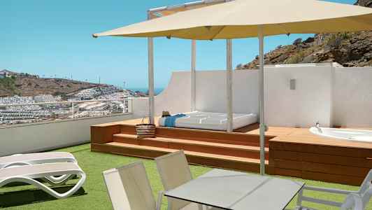 Terrasse med liggestole, bord, parasol og udsigt over et kuperet område med bygninger. (AI-genereret tekst)