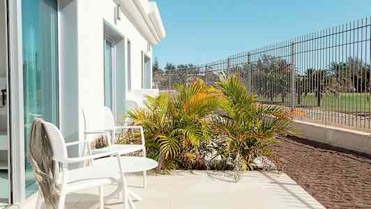 Terrasse med hvide stole og bord, omgivet af grøn vegetation og hegn. (AI-genereret tekst)