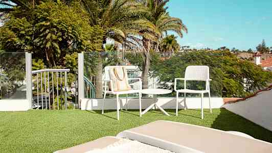 En terrasse med grøn syntetisk græs, møbler med hvide stole og et bord samt udsigt over frodige palmer mod en klar himmel. (AI-genereret tekst)