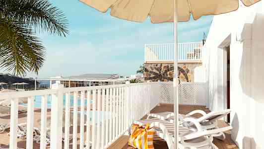 Udsigt over en terrasse med liggestole, parasol og en strandtaske. I baggrunden ses en pool og palmer. (AI-genereret tekst)