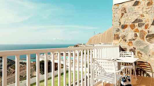 Udsigt fra en balkon med et bord og stole, og en udsigt over havet og omgivelser. (AI-genereret tekst)