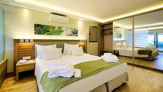 Et hotelværelse med en dobbeltseng, et grønt tæppe og hvide puder samt et spejl og garderobe i baggrunden. (AI-genereret tekst)