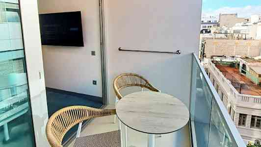 Billede af en altan med to stole og et bord samt udsigt over byens bygninger. (AI-genereret tekst)