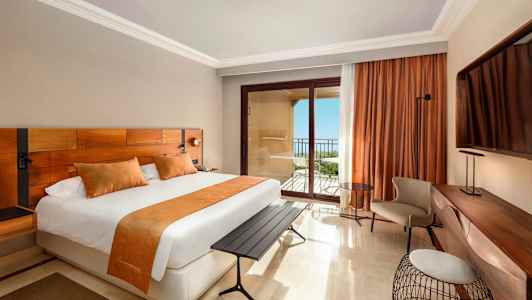 Et hotelværelse med en stor seng, orange indretningsdetaljer, et skrivebord og en altan med havudsigt. (AI-genereret tekst)