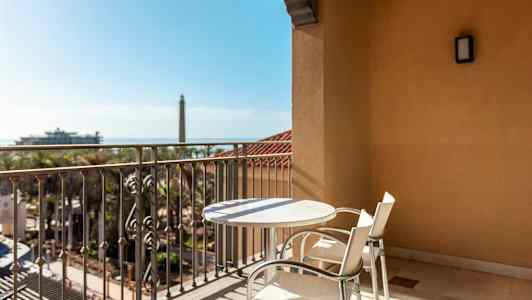 En balkon med et bord og to stole, udsigt over et landskab med palmer og et fjernt fyrtårn. (AI-genereret tekst)