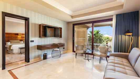 Et hotelværelse med moderne indretning, herunder siddeområde og sofa, et TV monteret på væggen, og store glasdøre, der fører ud til en balkon med udsigt over palmer. (AI-genereret tekst)