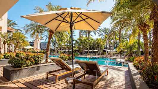 Et poolområde med to liggestole under en parasol, omgivet af palmer og blomster. (AI-genereret tekst)