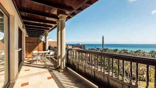 En solrig altan med bord og stole, trædetaljer og udsigt over havet samt et fyrtårn. (AI-genereret tekst)