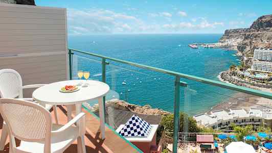 Udsigt fra altan med bord og to stole, hvor glas er serveret, mod havet og kystlandskab. (AI-genereret tekst)