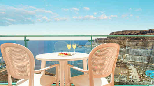 Billede af en altan med udsigt over havet og omkringliggende bygninger. På altanen står et bord med to glas og en tallerken med frugt samt to stole. (AI-genereret tekst)