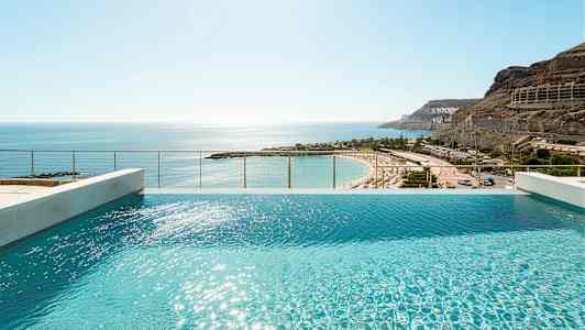 Udsigt over en infinitypool med solskin på vandoverfladen og en sandstrand i baggrunden. (AI-genereret tekst)