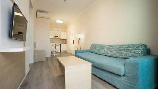 Et opholdsrum med en blå sofa og et bord foran, samt et TV på væggen og et integreret køkken i baggrunden. (AI-genereret tekst)