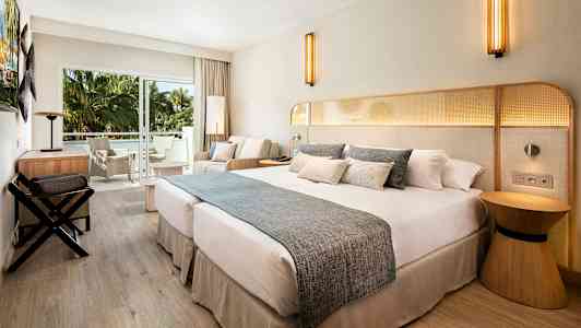 Et hotelværelse med en stor dobbeltseng, siddeområde og altan med udsigt til grønne palmer. (AI-genereret tekst)