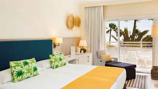 Et hotelværelse med en dobbeltseng med gult sengetæppe og grønne pyntepuder, lys indretning samt en balkon med udsigt over palmer og havet. (AI-genereret tekst)