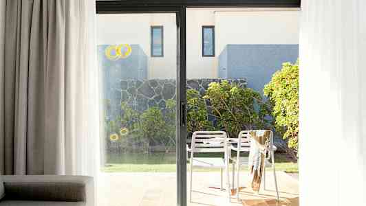 Billede af en terrasse med to hvide stole og et bord, omgivet af grønne planter og en stenmur, set gennem en glasskydedør. (AI-genereret tekst)