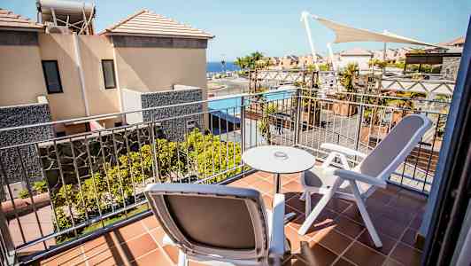 En altan med to stole og et bord, udsigt til bygninger og en pool. (AI-genereret tekst)