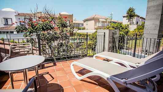 Billedet viser en balkon med et bord, to stole og to liggestole foran et rækværk, med udsigt over en indre gårdhave og omkringliggende bygninger. (AI-genereret tekst)