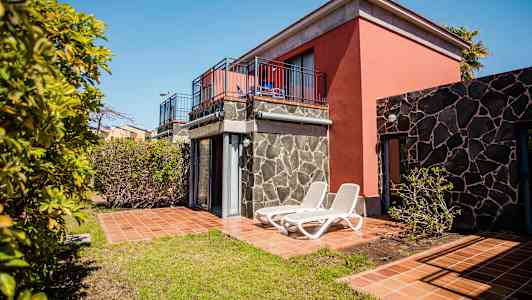 En ferielejlighed med terrasse og liggestole, omgivet af træer og grønne områder. (AI-genereret tekst)