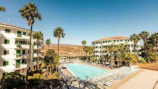 Billede af et hotelområde med en stor udendørs pool omgivet af solsenge. To hvide bygninger med grønne vinduer har palmer i forgrunden. (AI-genereret tekst)