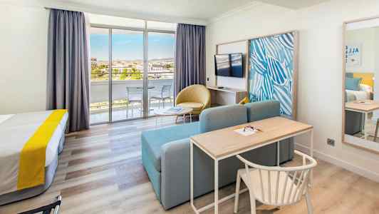 Et hotelværelse med seng, skrivebord, sofa, tv og glasdøre, der fører ud til en altan. (AI-genereret tekst)